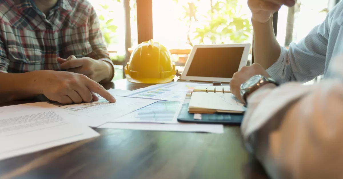 Two people sitting at a desk covered with scattered papers, a yellow construction hat, a tablet, and an open day planner.