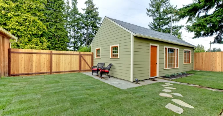 A small green dwelling unit set up in a fenced yard. It features an orange door and windows with orange and white trim.