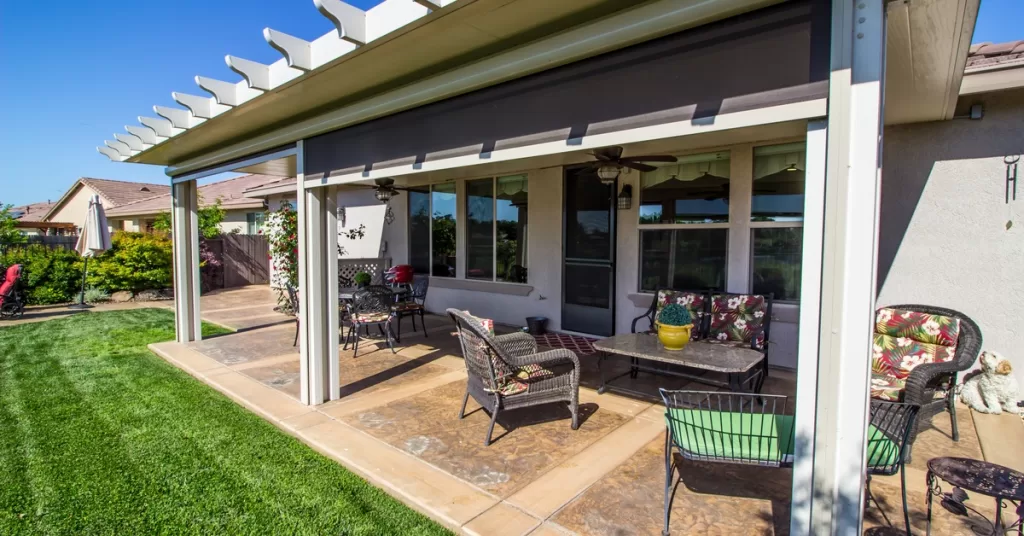 A covered patio features roller shades, black wicker furniture, and brown tile flooring beneath the seating area.