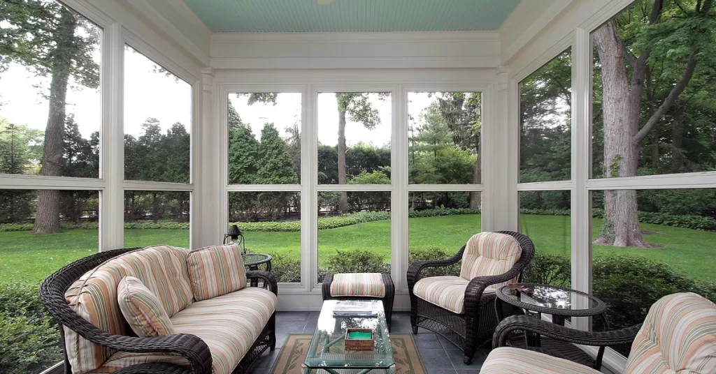 A white ceiling fan hangs from a turquoise ceiling in a screen room with white framing and black wicker furniture.