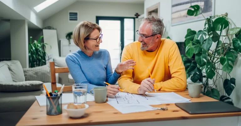 A woman and man sit at a wooden table with a blueprint spread out as they look at each other, both holding pencils.