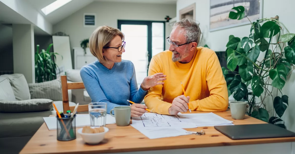 A woman and man sit at a wooden table with a blueprint spread out as they look at each other, both holding pencils.