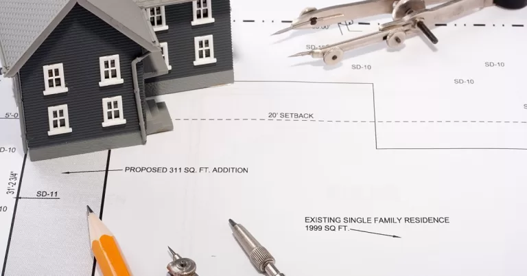 A model house, a pencil, and two measuring tools resting on top of a printed outline of a home addition.