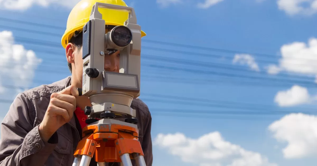 A person in a yellow hard hat peering through a theodolite. Blurry powerlines are visible in the background.