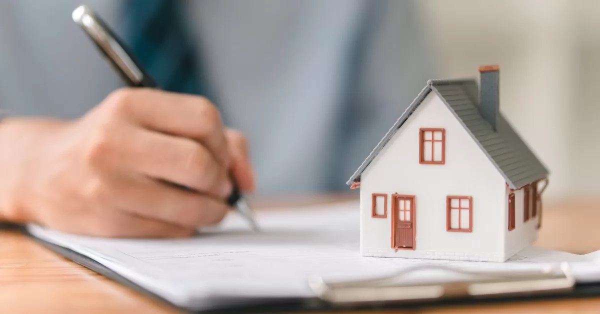A close-up of a hand with a pen writing on a clipboard topped with a white model house featuring a gray roof and chimney.