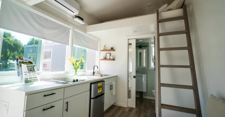 A compact dwelling interior featuring a kitchen, loft bed with ladder, open bathroom door, and fliers on a counter.