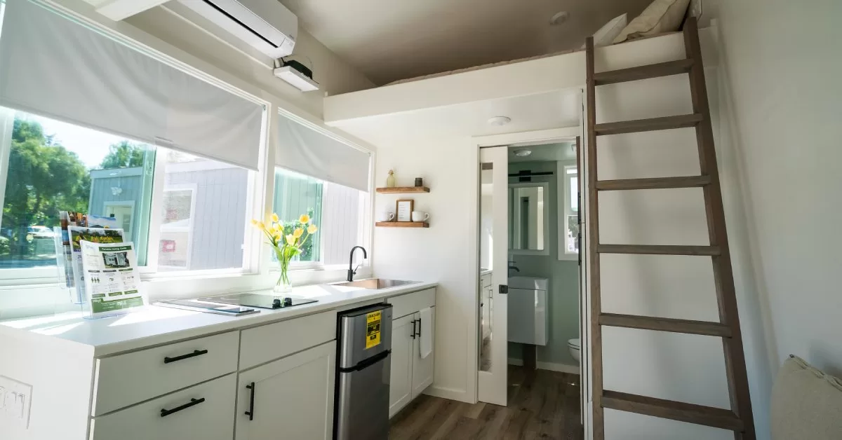 A compact dwelling interior featuring a kitchen, loft bed with ladder, open bathroom door, and fliers on a counter.