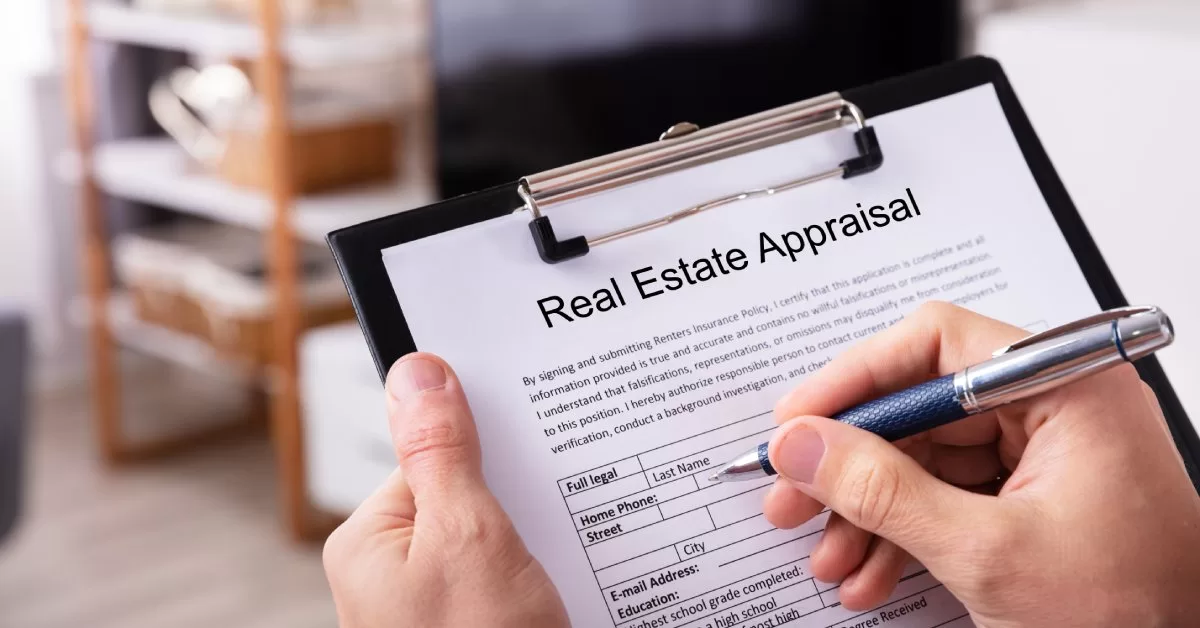 A close-up of a hand with a pen resting on a clipboard with a paper labeled "Real Estate Appraisal."