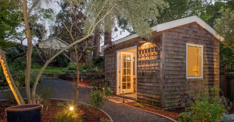 A warm light glows from a timber unit with open white doors, as a tree stands across a pathway from the entrance.