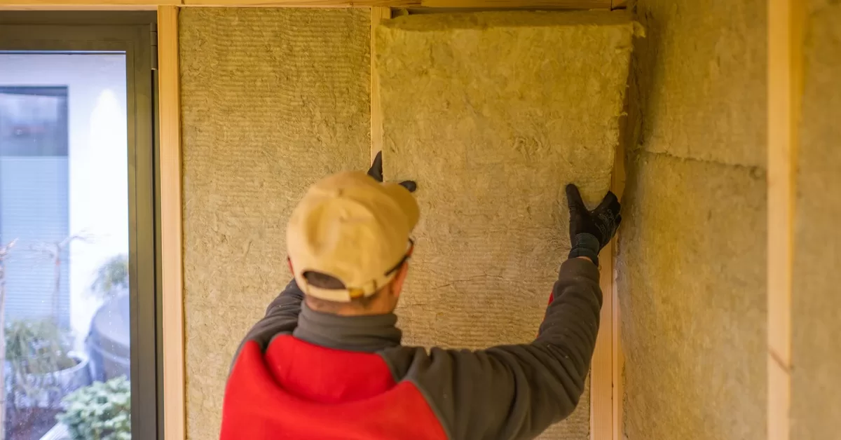 A man in gloves and a cap places insulation on a wall between wooden framing near a glass door.