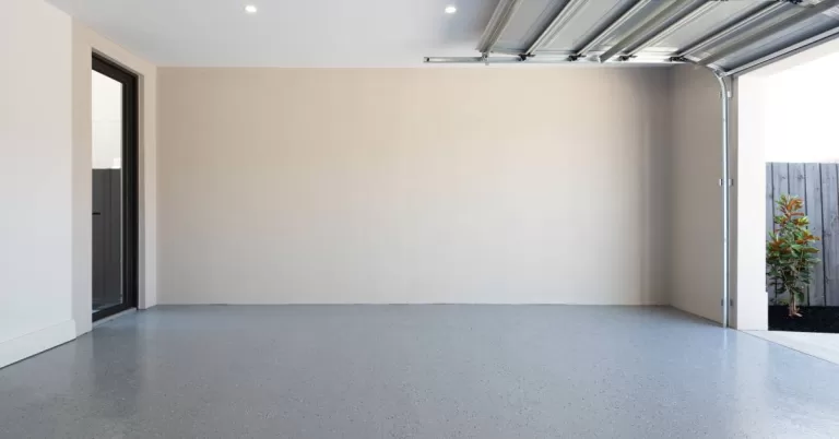 An open garage door lets natural light flood an empty beige garage with speckled gray flooring and a white ceiling.