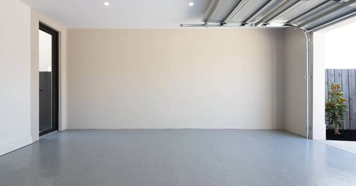 An open garage door lets natural light flood an empty beige garage with speckled gray flooring and a white ceiling.