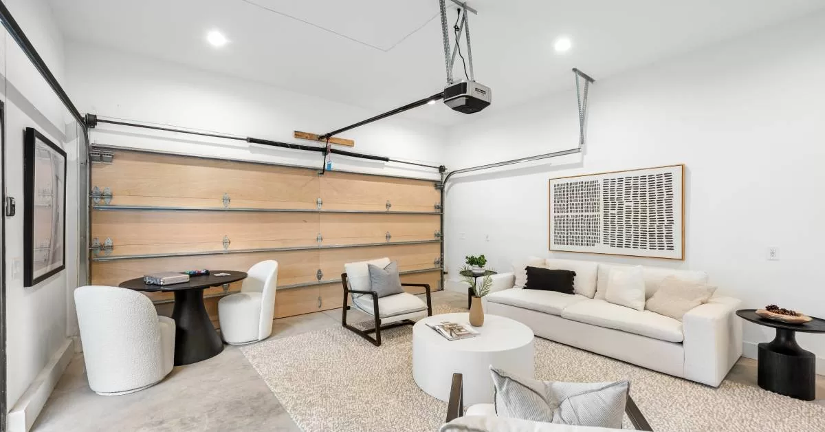 A garage interior features white walls, a pale wood garage door, a cream couch, gray flooring, and a beige rug.