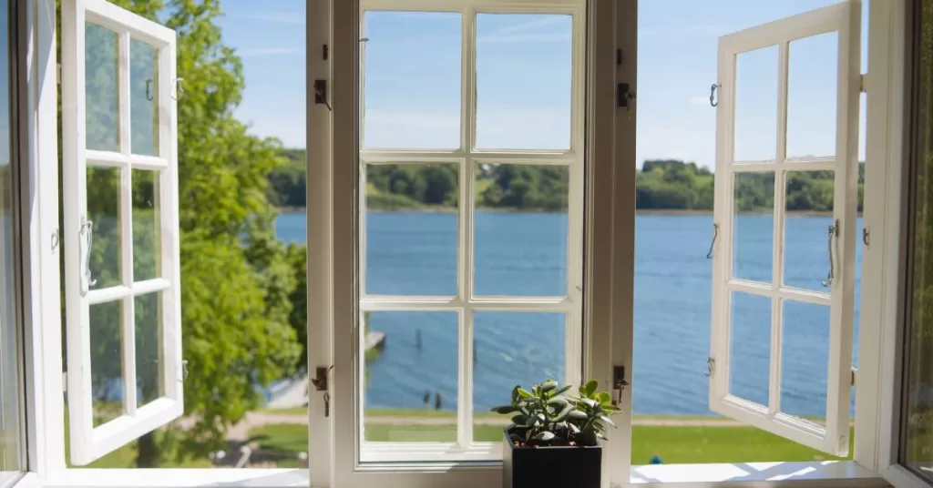 Open side windows frame a central window with a potted plant in front of it, offering a view of water and a dock behind.