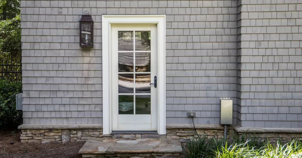 A white lited door with a black lantern-style light to its left is on a gray home with greenery near the path.