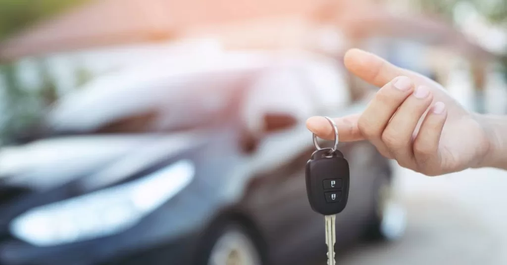 A hand holds a key with a black fob featuring two lock buttons, set against a black sedan and homes in the background.