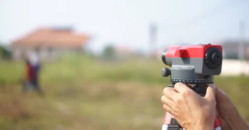 A pair of hands holds a red and charcoal total station, with green land and a beige building blurred in the background.