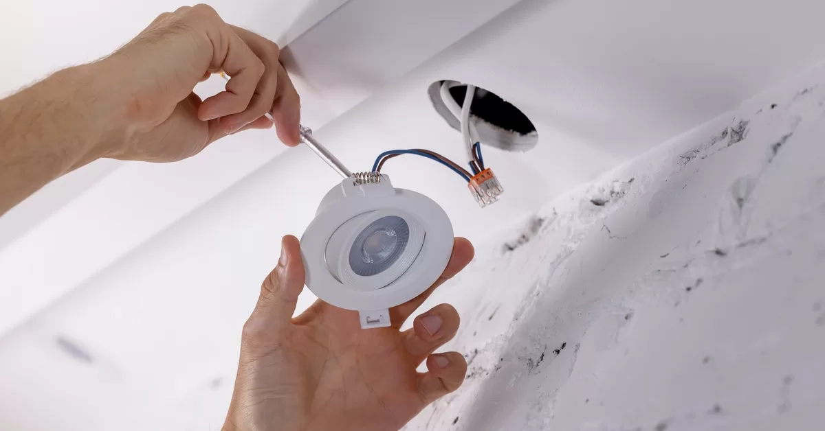 Bare hands secure a recessed LED light with a screwdriver while wiring extends from a drywall ceiling hole.