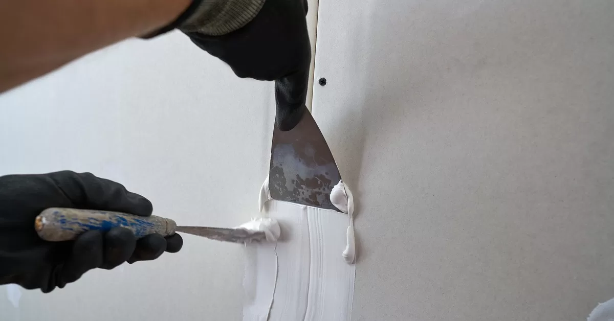 Gloved hands spread white plaster over light gray plasterboard seams using two different putty knives.