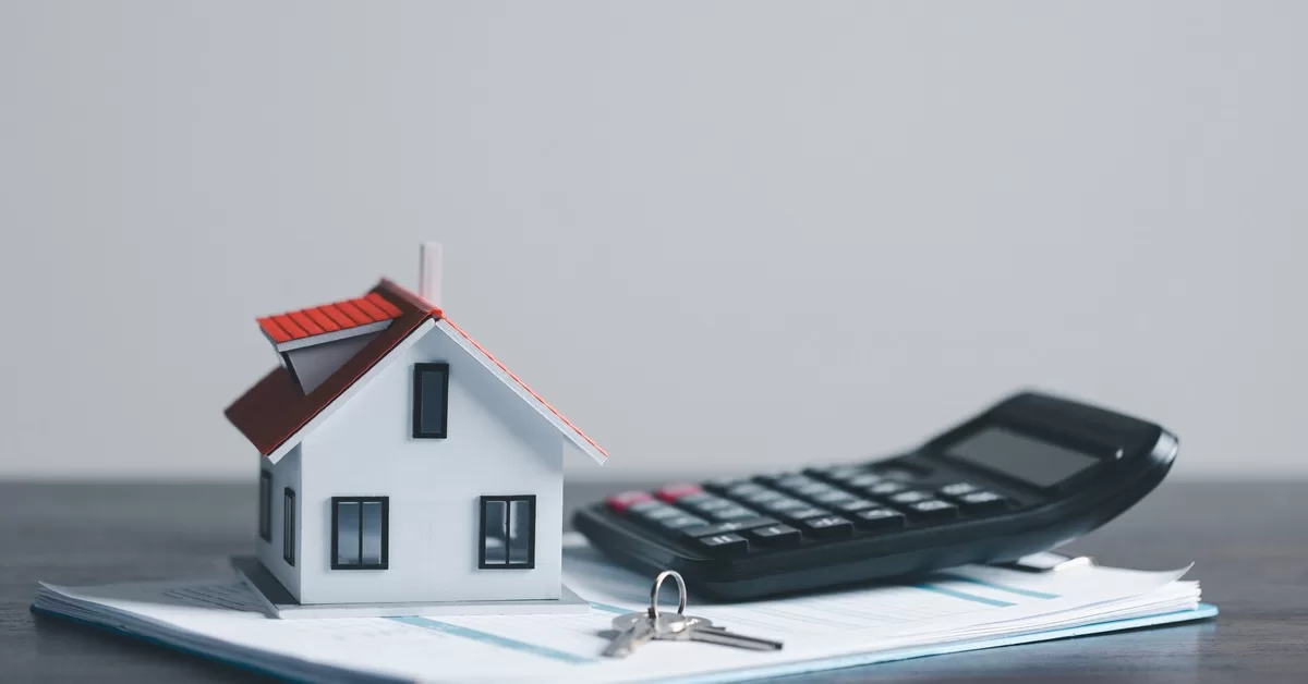 A white model house with a red roof, a pair of keys, and a calculator rest on top of documents on a gray table.