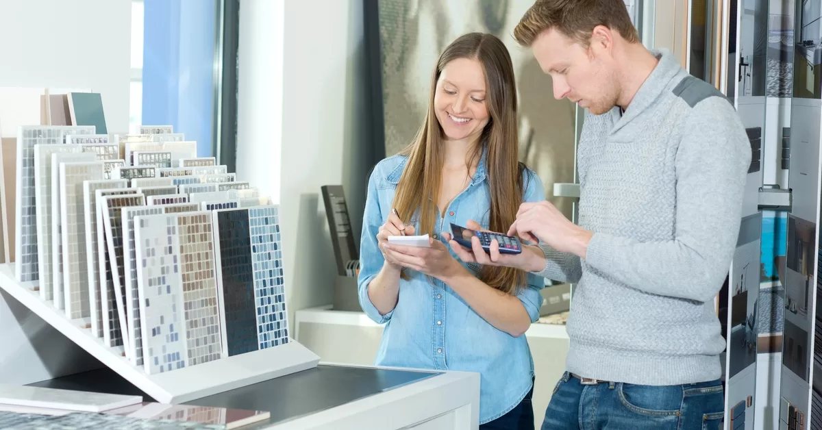 A woman writes on a notepad while a man uses a calculator. Tile samples are arranged across a counter in front of them.