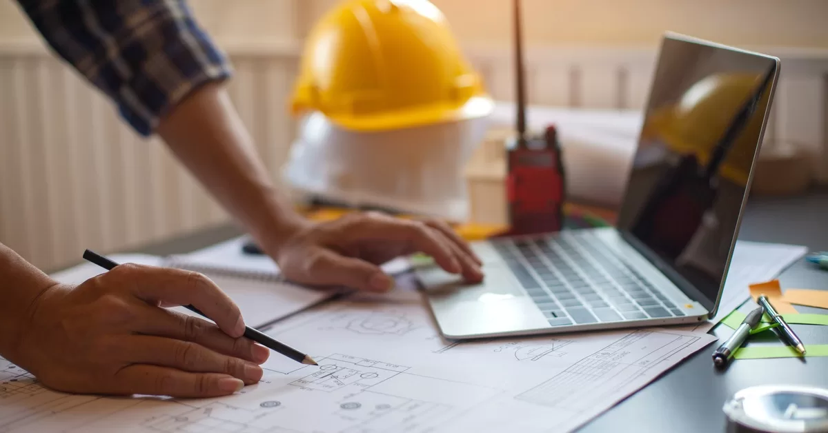 A person uses a laptop with one hand and holds a pencil above a blueprint with the other. A hard hat sits in the background.