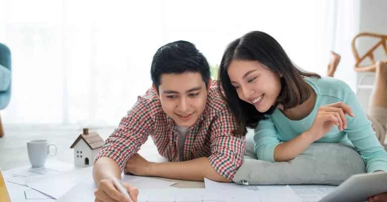 A man and woman lie on a gray carpet while reviewing a blueprint. A white model house and a mug sit beside the man.