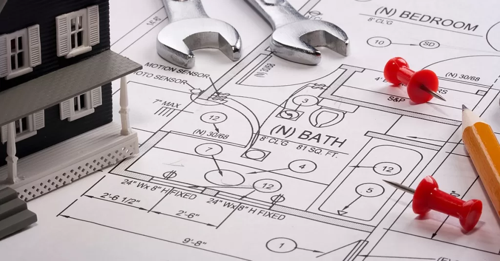A pencil rests on schematics showing a bathroom and a bedroom. Two red pushpins, two wrenches, and a black model house lie nearby.