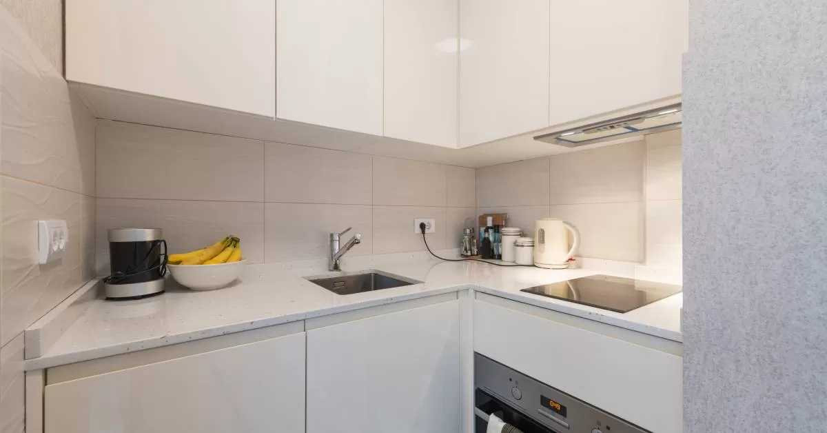 White cabinets sit above and below large rectangular white tiles. A kettle sits between a small sink and a stovetop.