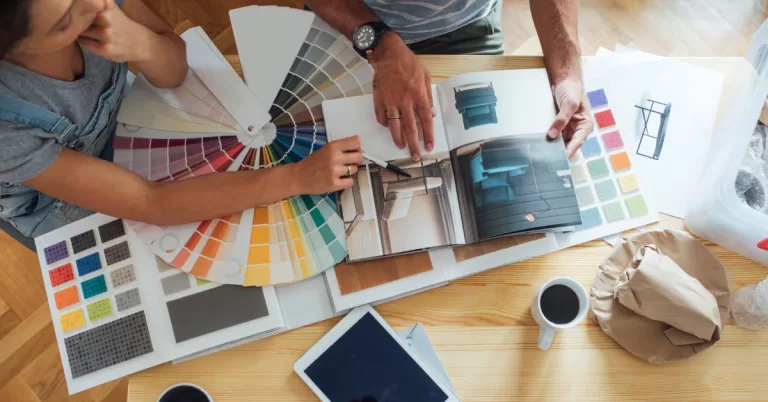 Two people examine a furniture catalog on a table covered with paint and flooring swatches.