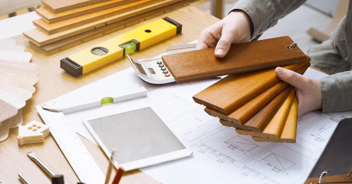 A pair of bare hands holds wooden baseboard samples over a house blueprint surrounded by tools, screws, and a tablet.
