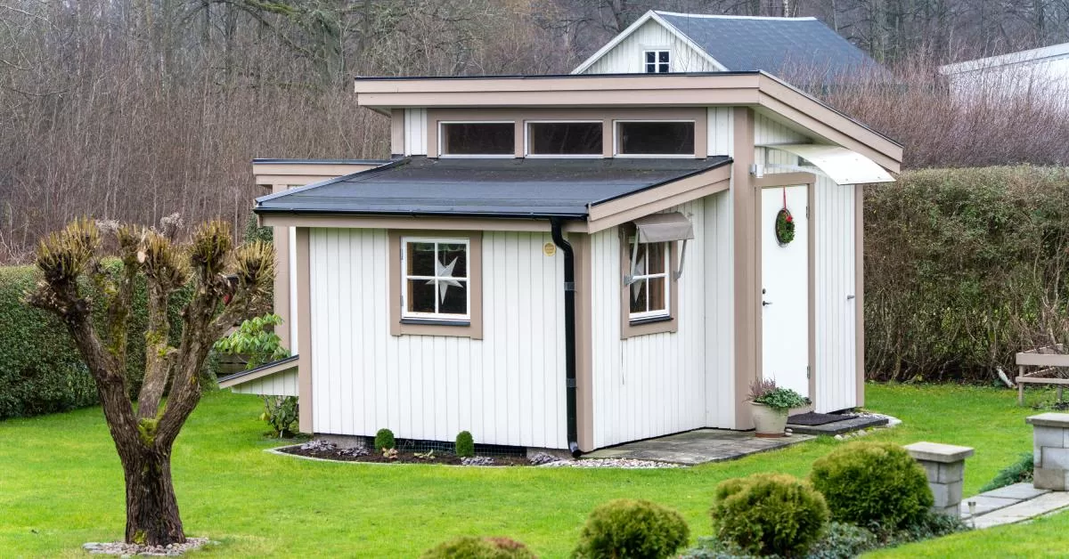 A small white ADU with a black roof features a wreath on the door, silver stars in the windows, and trimmed shrubs.
