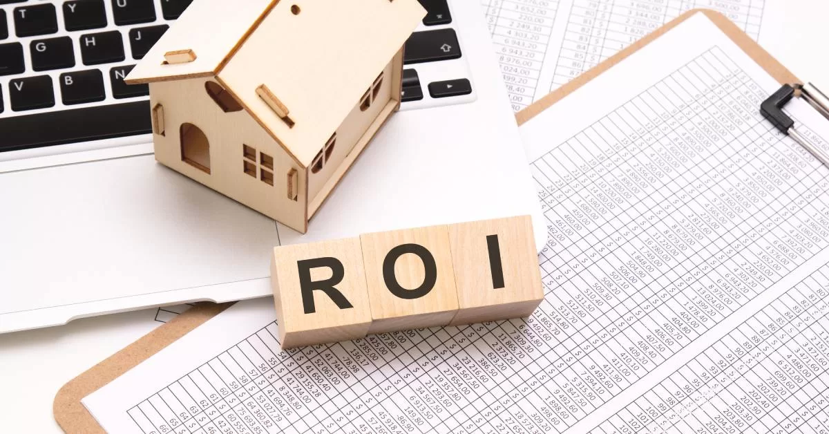 A wooden house figurine rests on a laptop touchpad above printed data tables and wooden cubes spelling “ROI”.