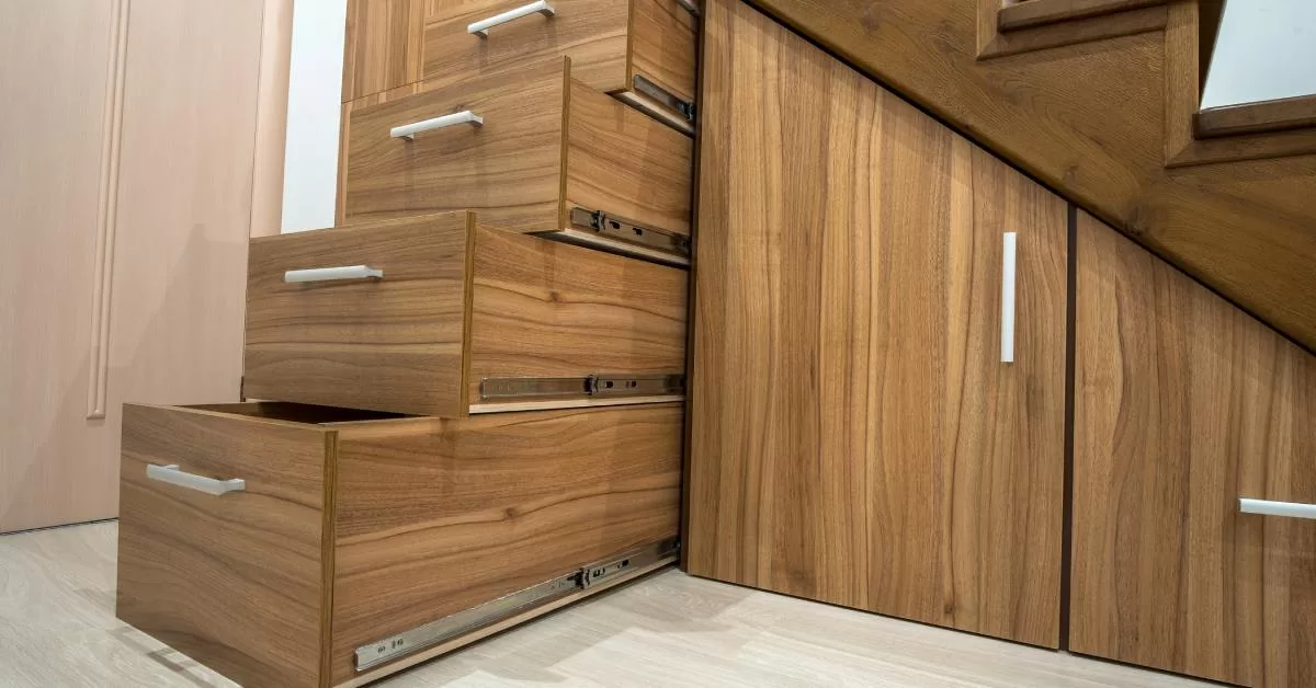 Five white-handled wooden cabinets slide out from under glossy wooden stairs over gray laminate flooring.