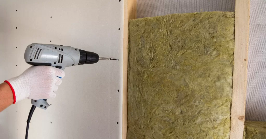 A person uses a gray electric screwdriver to drive a screw into drywall near timber and mineral rock insulation.
