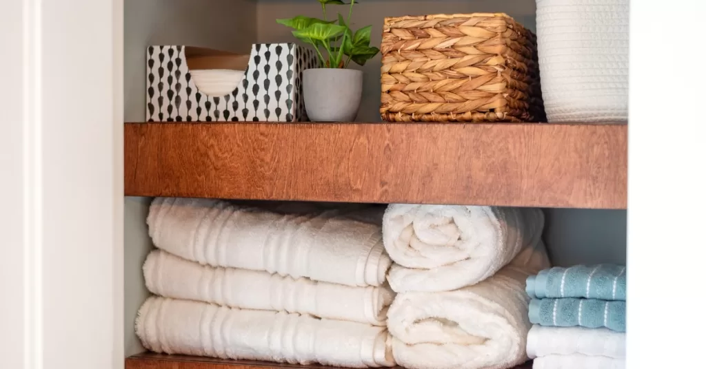 Towels, a tissue box, a potted plant, and a wicker container sit on two wooden shelves against gray closet walls.