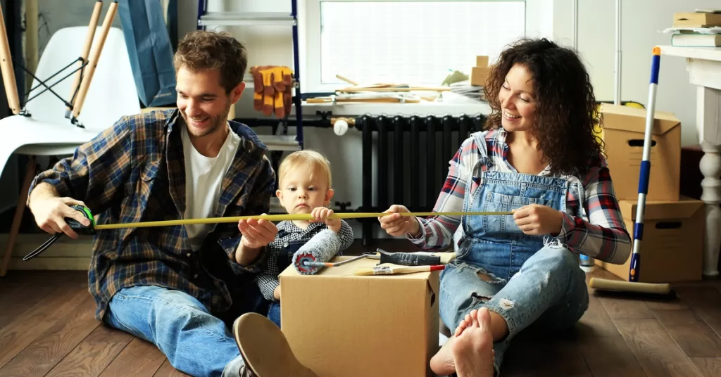 A man, a woman, and a child hold a measuring tape near a box with a paintbrush and roller, surrounded by boxes and luggage.