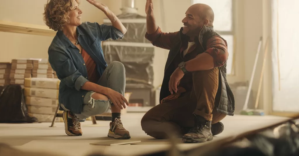 A man and a woman squat with their hands raised. Behind them is a covered fireplace, surrounded by stacks of materials.