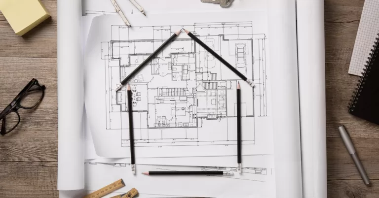 Black pencils form a house shape on top of blueprints, beside a pen, keys, Post-it notes, glasses, and a measuring tool.