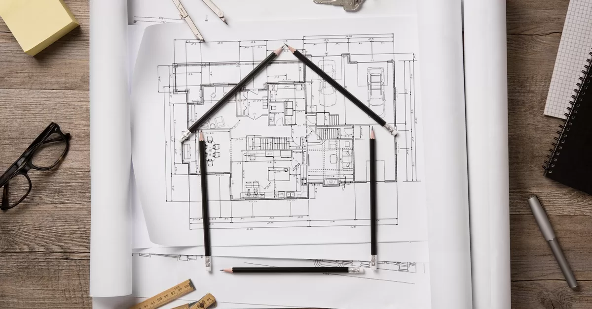 Black pencils form a house shape on top of blueprints, beside a pen, keys, Post-it notes, glasses, and a measuring tool.