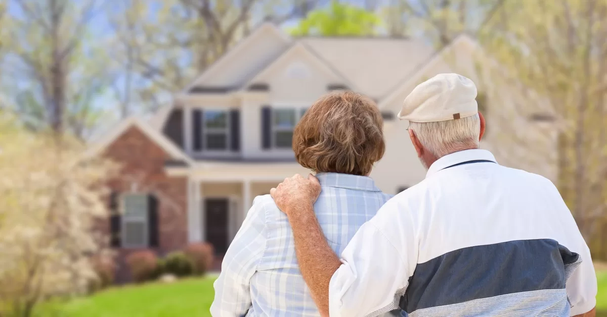 The back of an older couple standing next to each other and looking at their home. The man has his hand on the woman's shoulder.