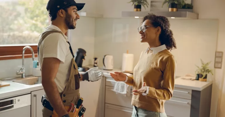 A handyman in overalls and a tool belt chats with a smiling woman in a modern, well-lit kitchen with light cabinets.