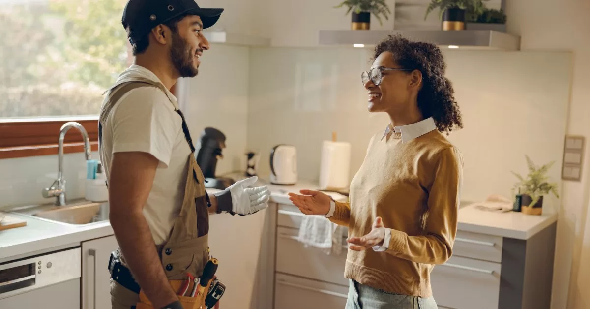 A handyman in overalls and a tool belt chats with a smiling woman in a modern, well-lit kitchen with light cabinets.