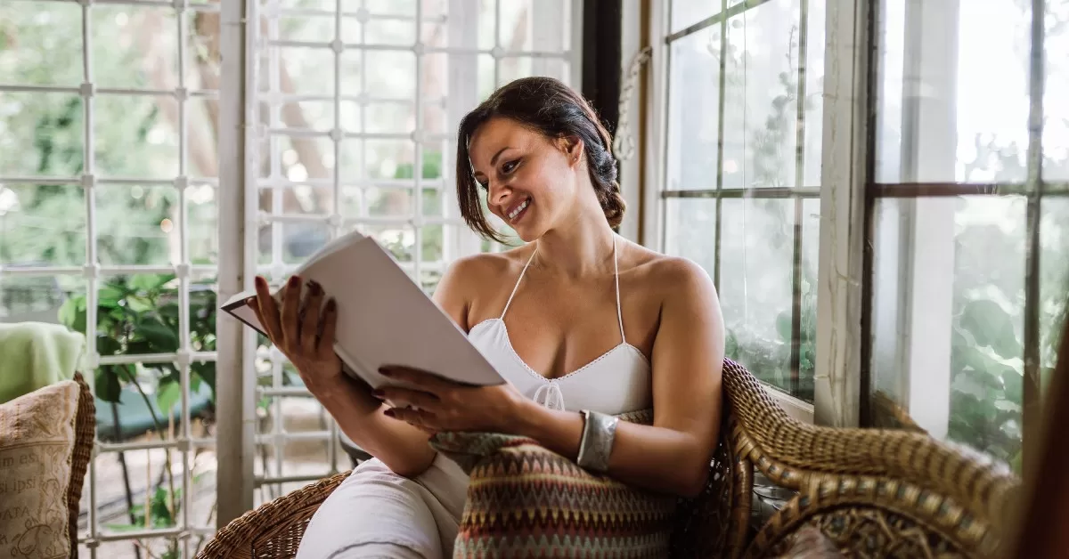 A woman wearing a white top and holding a white book sits on a couch and reads in her brand-new sunroom addition.