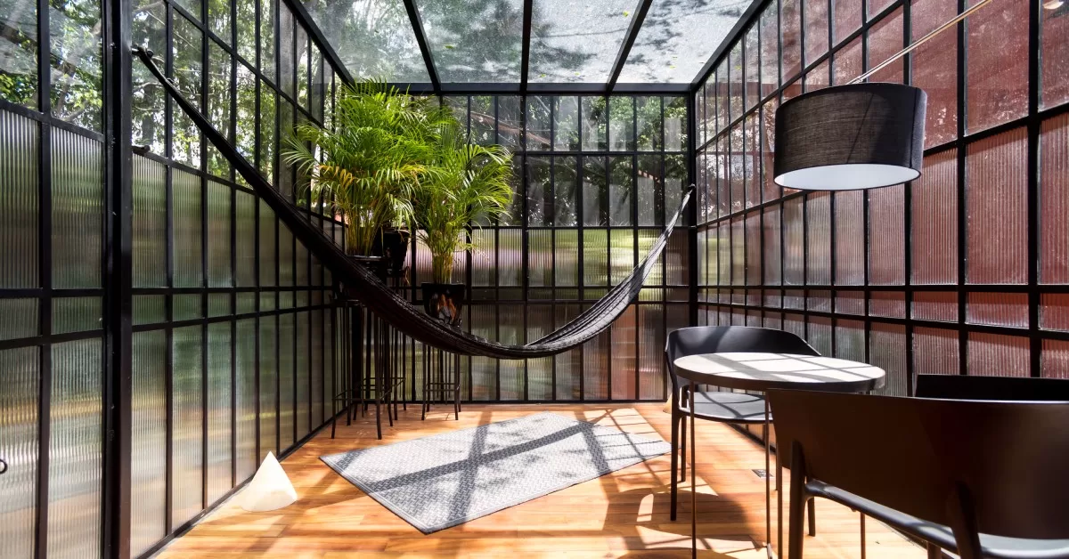A modern sunroom contains a glass ceiling, a hammock, potted plants, and a wooden floor, with natural sunlight streaming in.