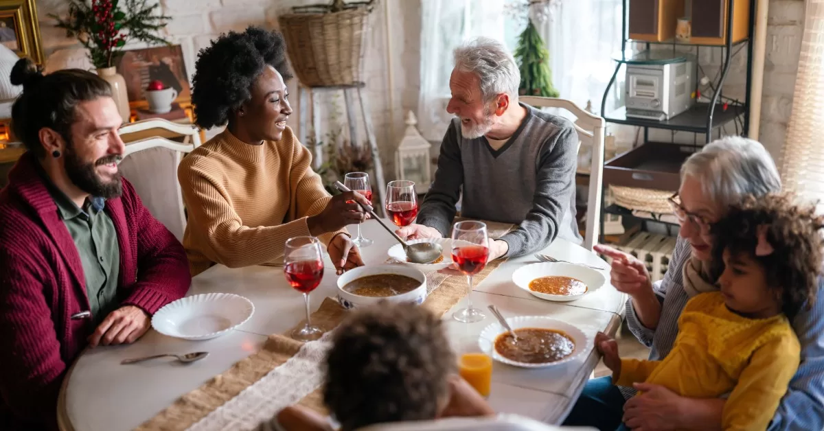 A diverse family shares a meal at a dining table in a bright, cozy room. There's food, drinks, and decorations visible.