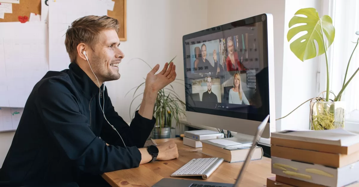 A man sits in a home office during a video call, with a monitor, laptop, books, and plants on his desk.