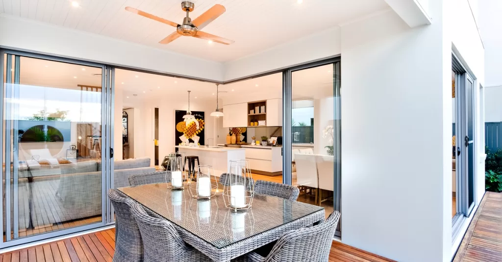 A modern outdoor deck contains a dining set, ceiling fan, and sliding glass doors connecting to a kitchen and living room.