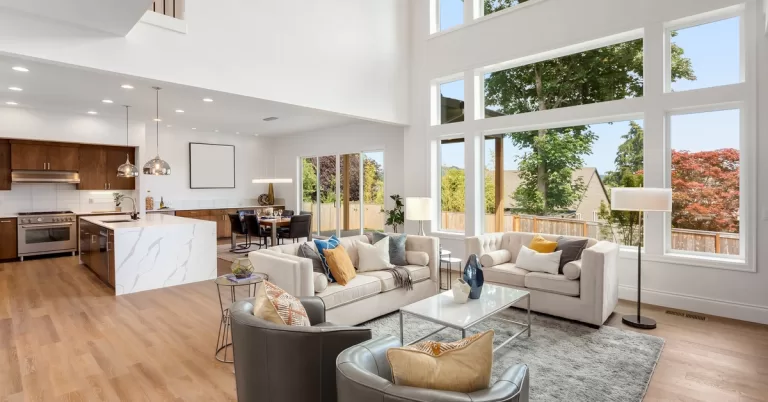 An open-concept home features a modern kitchen, dining area, and living room with high ceilings and large windows.