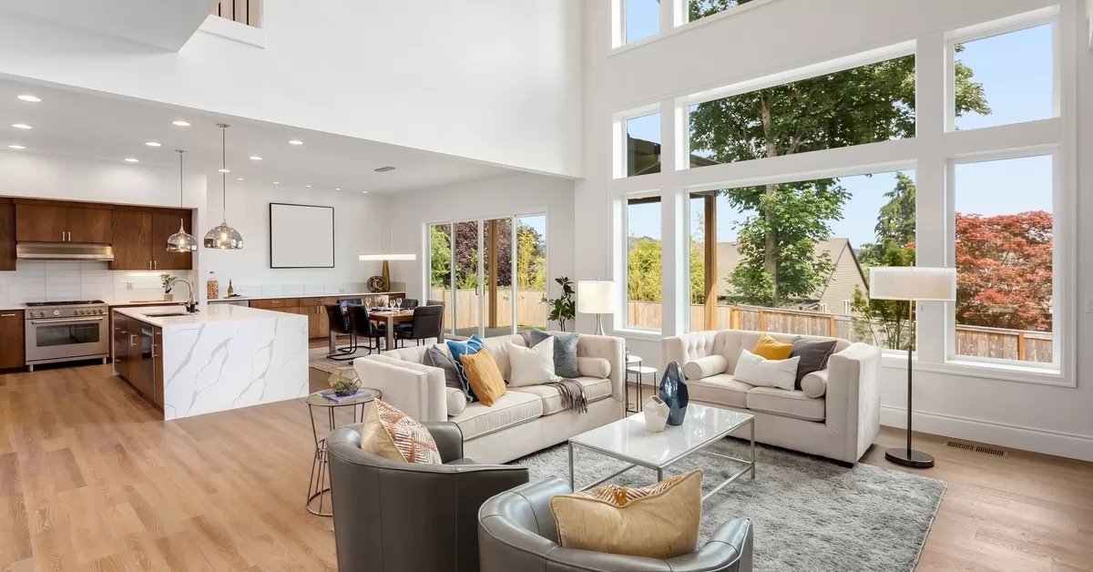 An open-concept home features a modern kitchen, dining area, and living room with high ceilings and large windows.
